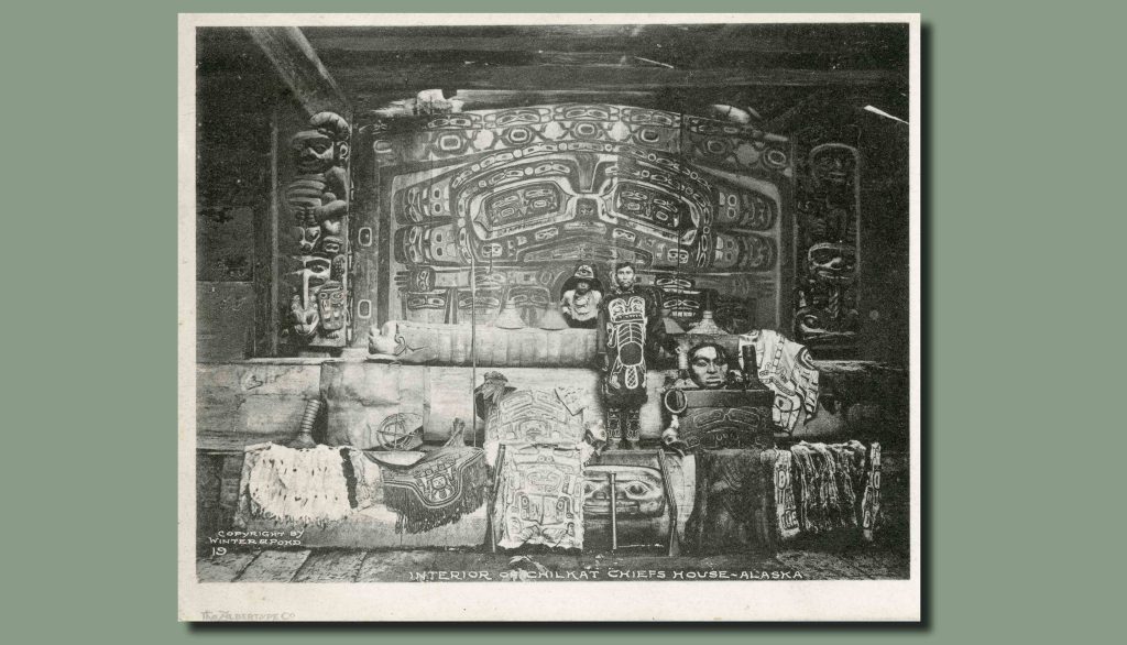 Black and white image of a man standing in front of a highly-decorated wall surrounded by sculptures, masks, robes, and blankets in the Northwest Coast style. A small child wearing a hat peers through a hole in the decorated wall. 