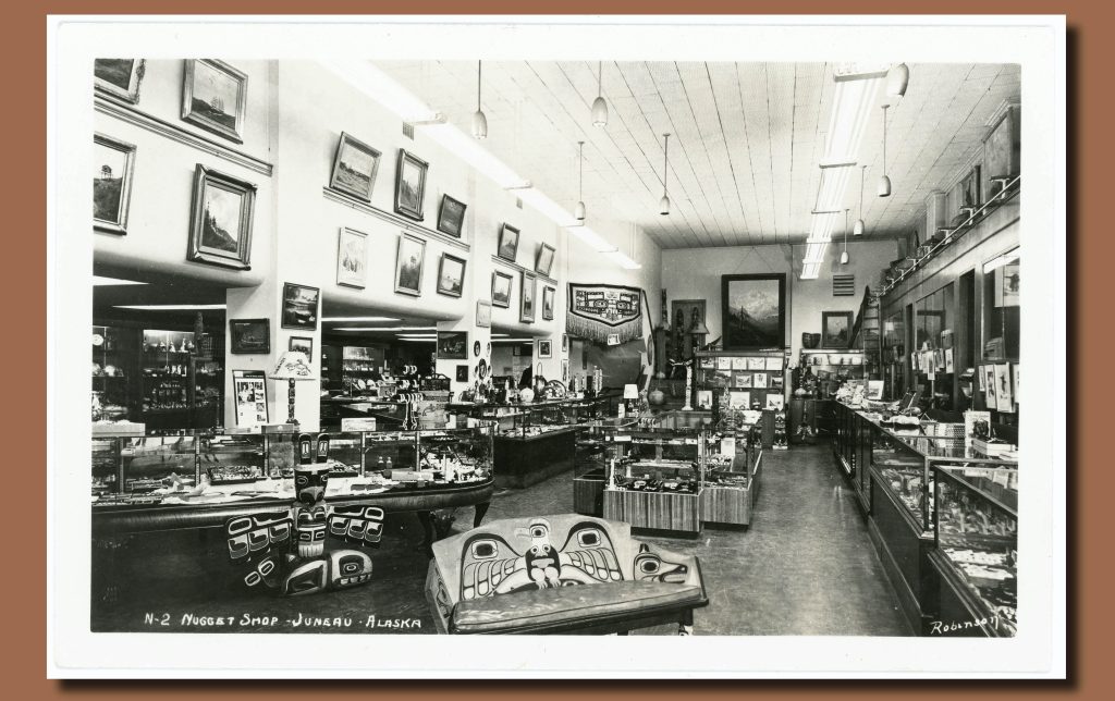 Black and white image of a store interior. Objects in the Northwest Coast style fill cases and the sales floor and framed pictures hang on a high wall.