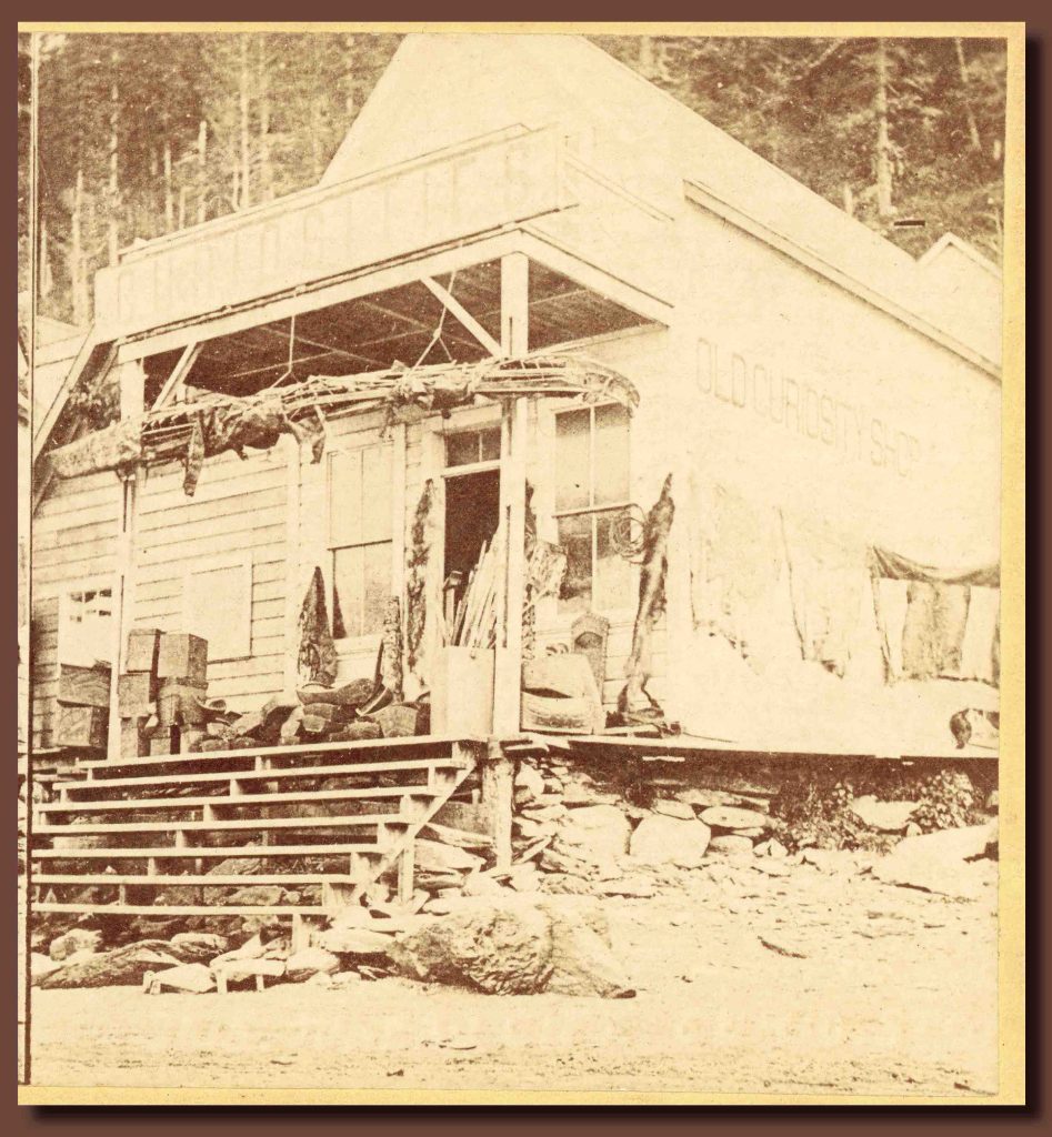 Sepia-tone image of a wooden buulding with steps leading up to a porch stacked with boxes and other items. A canoe or kayak shell hangs above the steps.