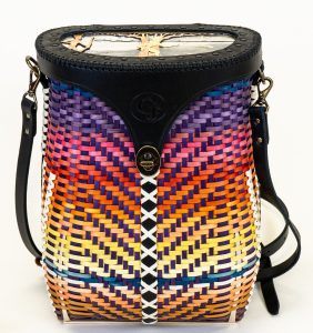 Image of a basket in the shape of a pack woven to look like a sunset over water. The cover is black leather with an inset metal scene of a man with a bow and another man emerging from a tree.