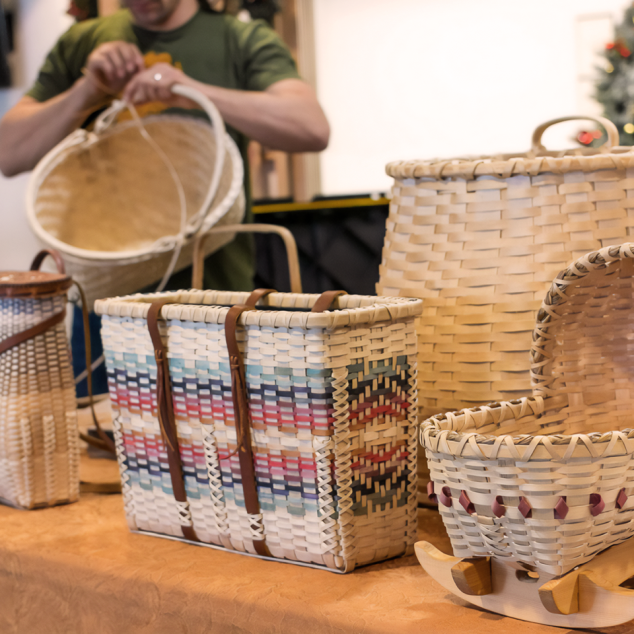 Image of baskets on a table. One is a small rocking cradle. One is a decorated in patterns of red and blue. One is a pack basket. A man works on a basket in the background.