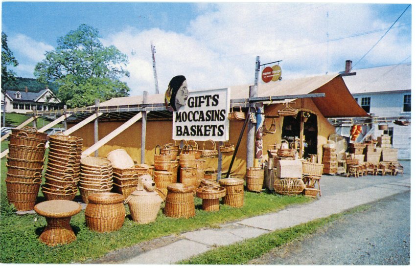 Image of a canvas tent surrounded by piles of baskets. A sign reads "Gifts Moccasins Baskets".