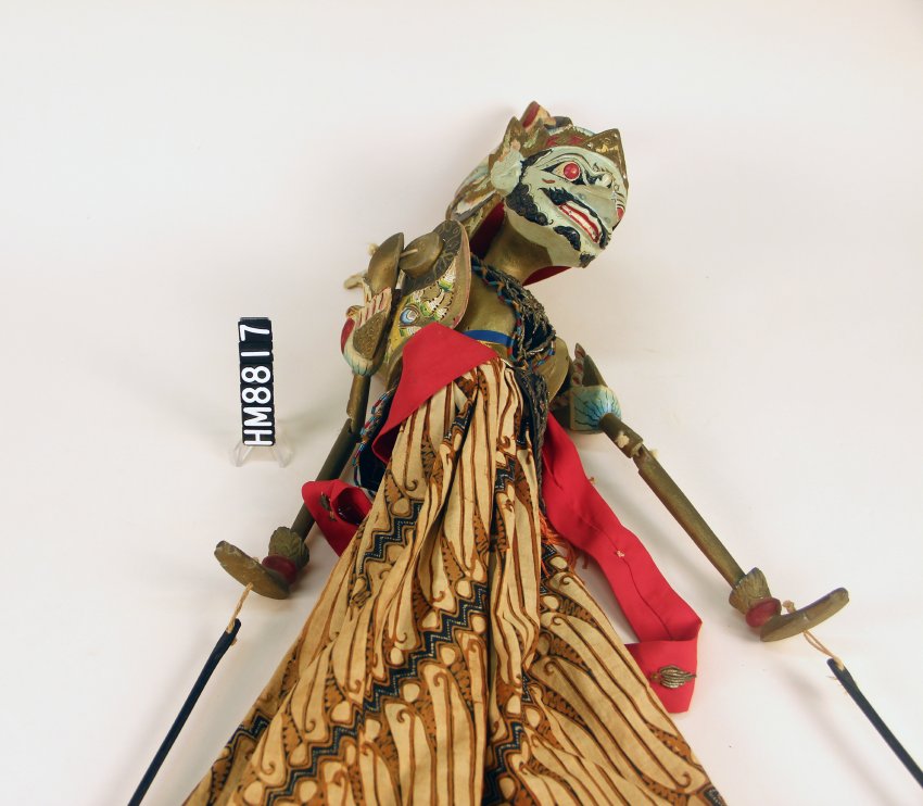 Image of a puppet with green skin and rich textiles. The hands are attached to sticks.