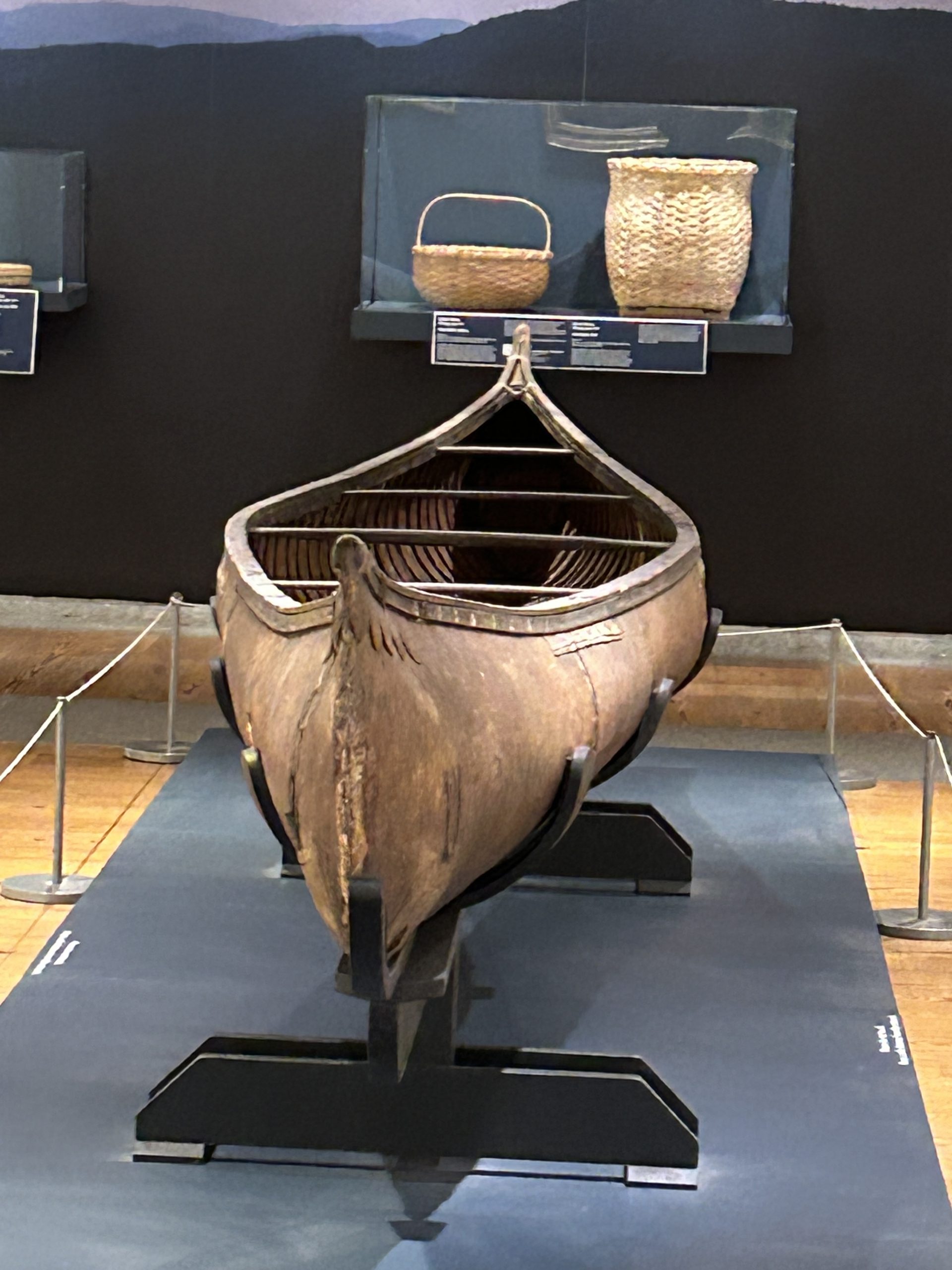 Image of a birchbark canoe in a museum exhibit with baskets in a case behind it.