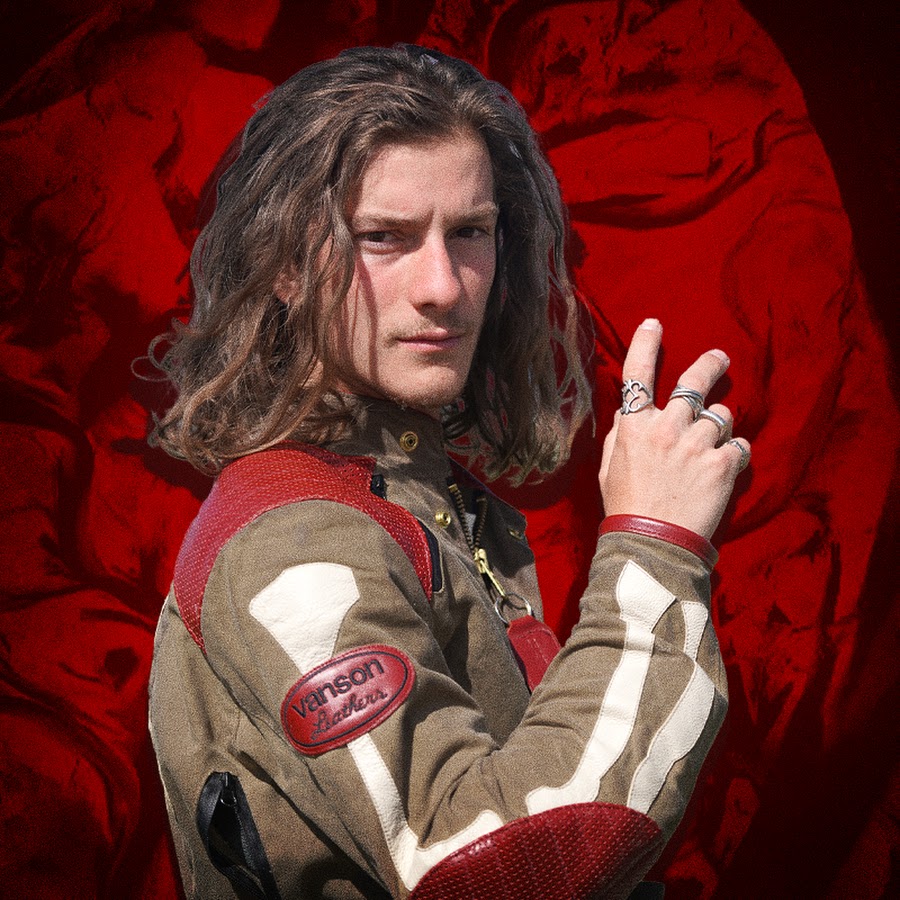 Image of Caucasian male with shoulder-length brown hair in front of a red background.