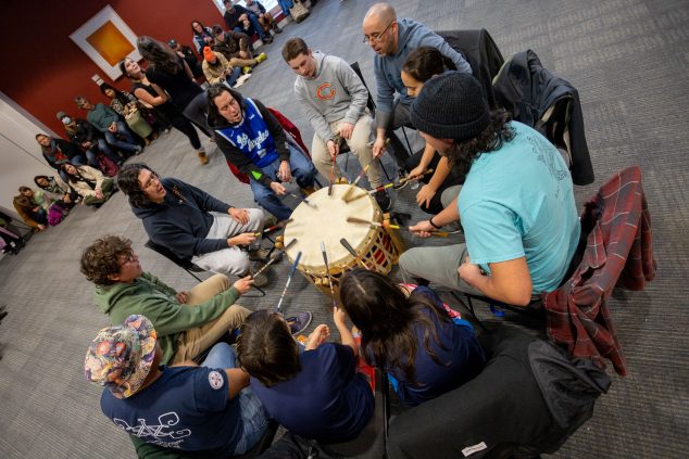 Image of a group of people sitting around a large drum, all holding drumsticks and drumming.