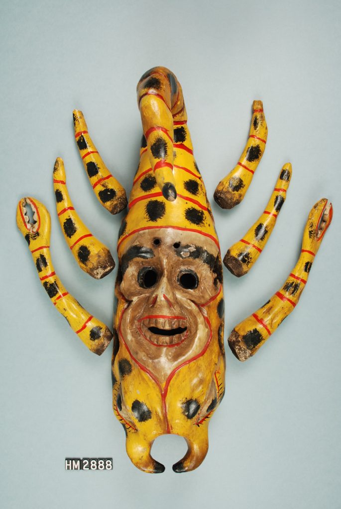 Image of a brightly-painted wooden mask in the form of a scorpion. A face has been carved on the back of the scorpion, which matches up with the wearer, and the tail of the scorpion sticks up above the face. Six "legs" are separate pieces that slot into holes on the sides of the mask.