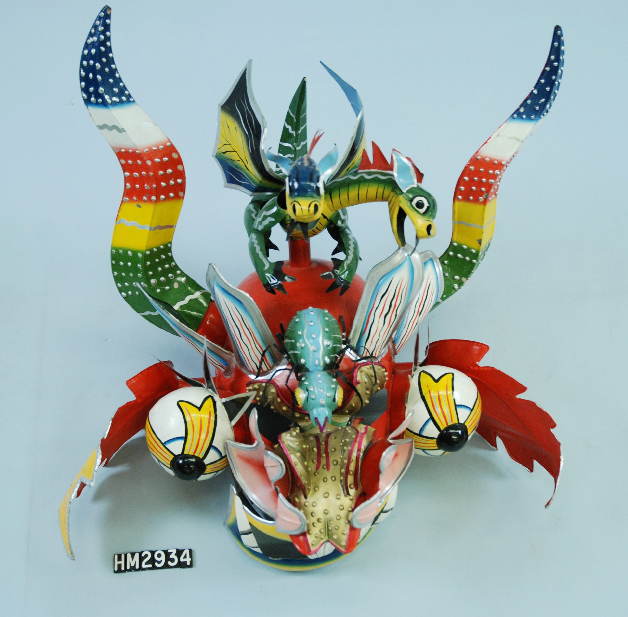 Image of a colorful metal mask with the face of a dragon, horns, smaller dragons on the top, and a large spider between the eyes.