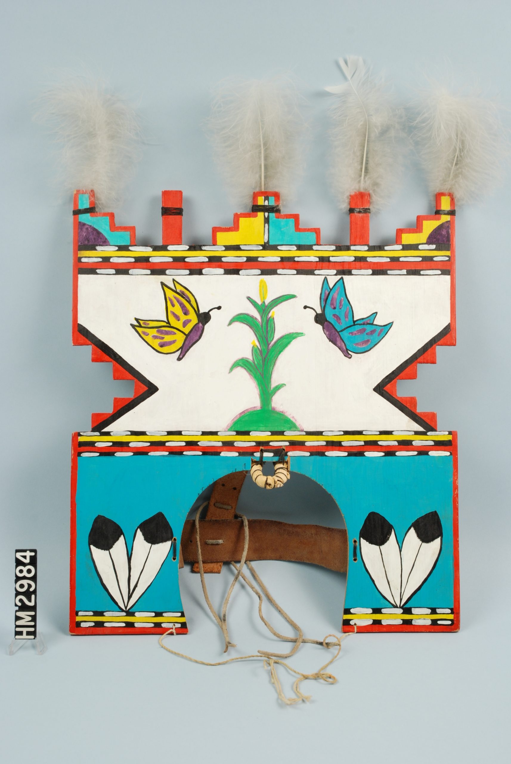 Image of a brightly-painted, flat wooden headdress with feathers at the top. It features corn and two butterflies.