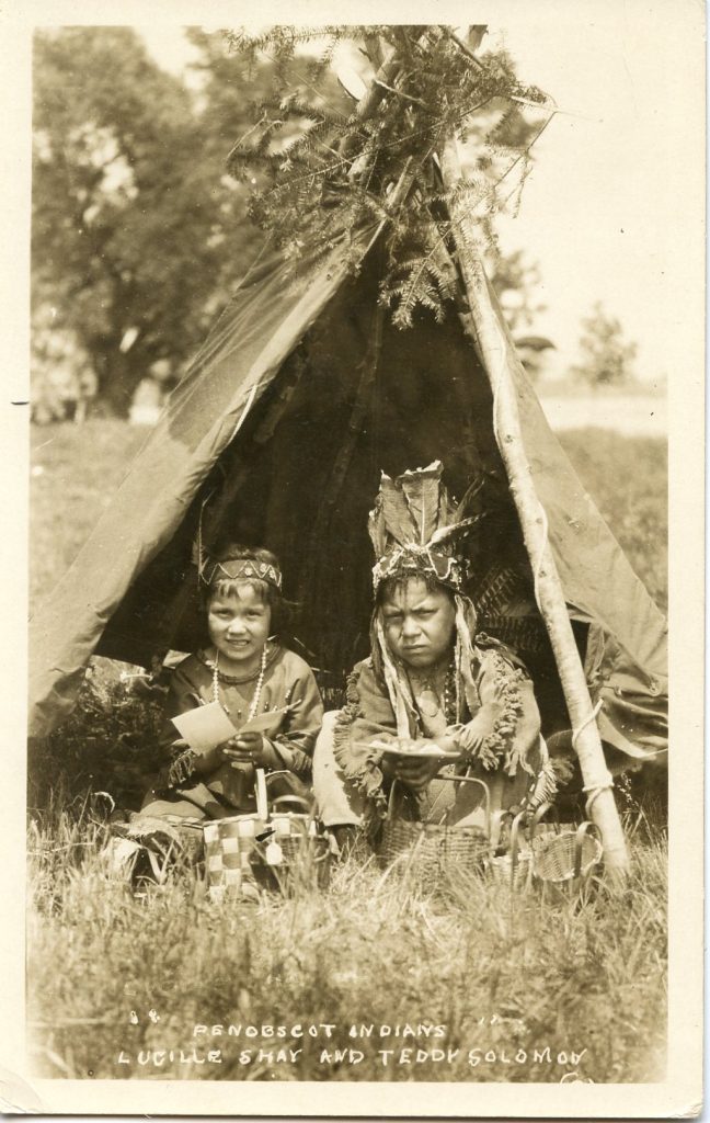 A sepia-tone image of two children sitting in a tiny tepee. They are wearing native dress. The girl wears a beaded headband and the boy a feathered and beaded headdress. The have baskets around them.