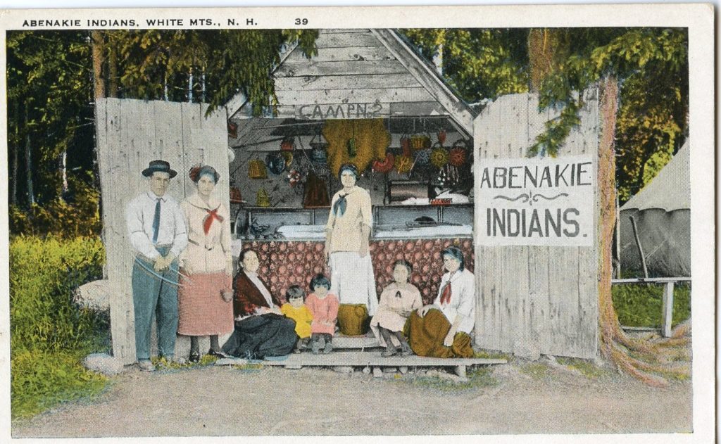 A colorized image of a group of people by a small wooden building, open to display baskets for sale. A sign reads "Abenakie Indians"