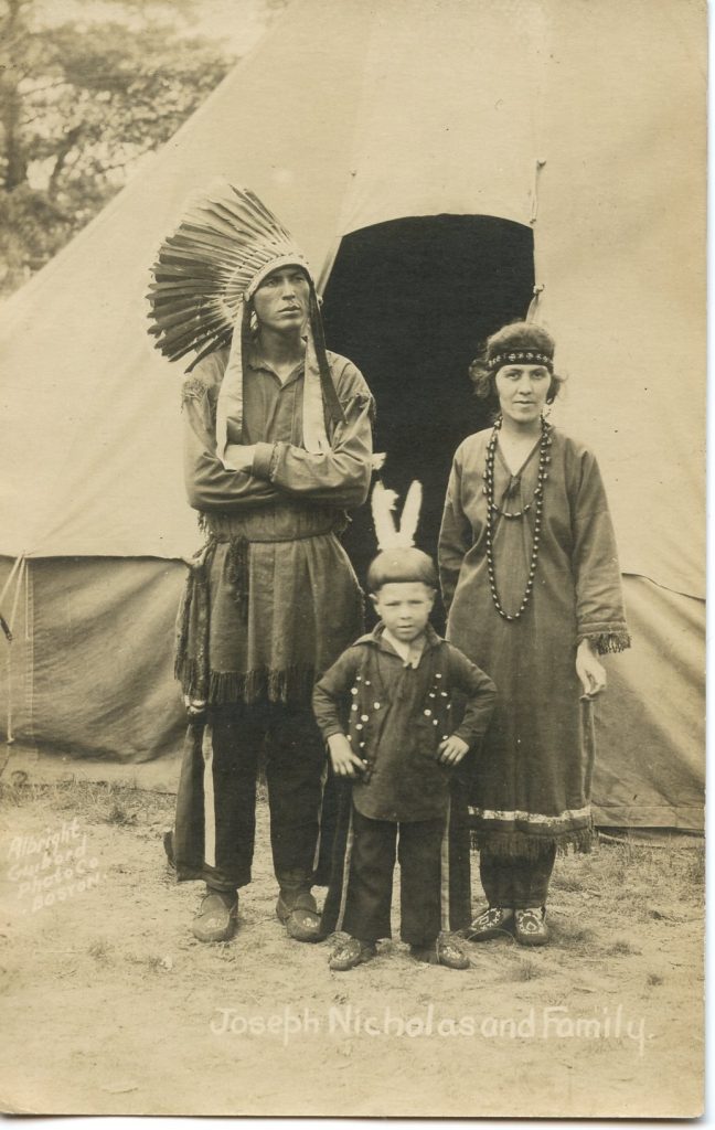 A sepia-toned image of a man, woman, and child standing in front of a tent. They all wear Native-style dress. The man is wearing a Plains-style feathered headdress, the woman a beaded headband with a single feather, and the small child two feathers.