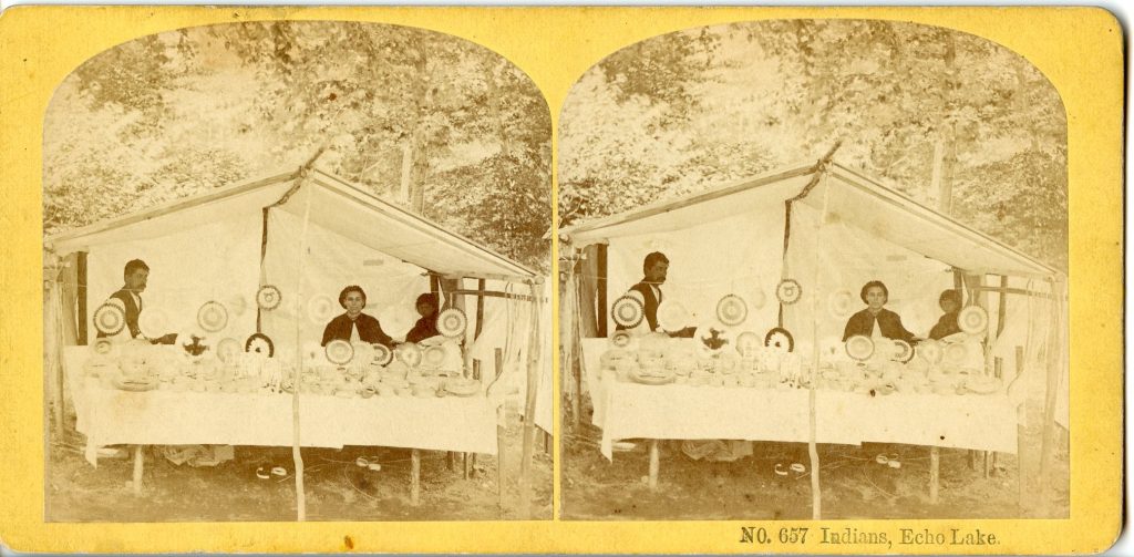 Stereograph of a grayscale image of three people inside a canvas tent behind a display of baskets on a table. Beneath the table can be seen rolls of brown ash weavers, suggesting they are making baskets.