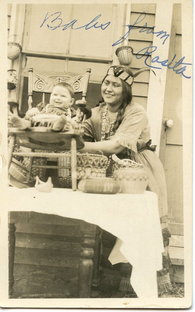 A sepia-tone image of a smiling woman in Native dress with a beaded headdress standing behind a table. On the table are a variety of baskets and a small carved chair in which she supports a smiling, chubby baby.