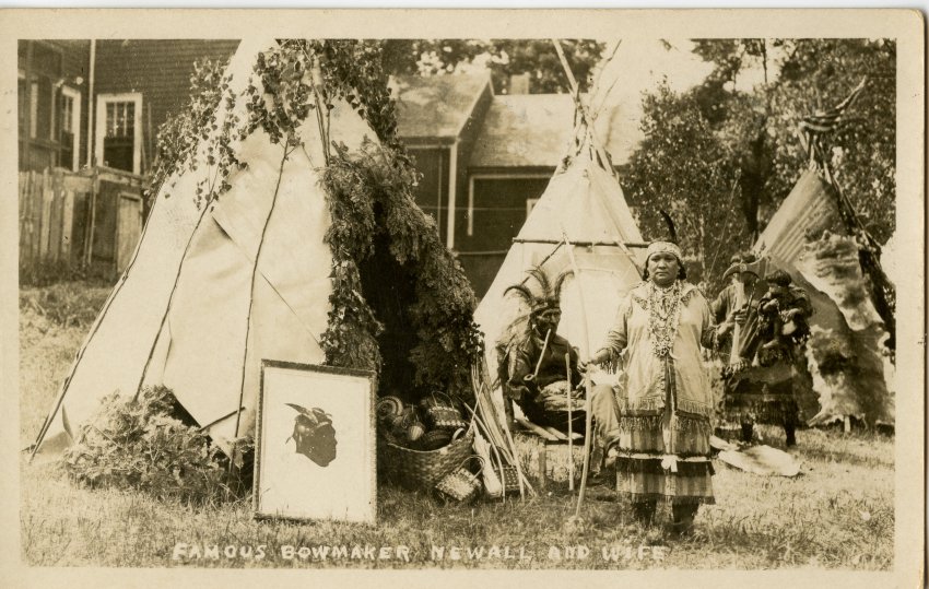 Sepia tone image of three tepees set up in front of houses. A woman in Native American dress stands holding a bow and hatchet. Behind her an elder in a feather headdress smokes a long pipe. A woman holds a child in the background.