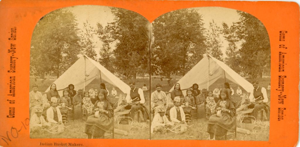 Stereoview of a grayscale image of a group of people sitting in front of a canvas tent. They were a mix of Western and Native dress, including beaded caps. Many of them are working on partially-finished baskets.