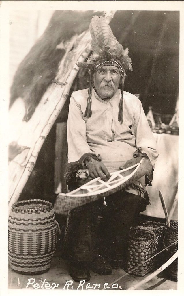 A grayscale image of a male elder sitting at the entrance of a tepee and smiling at the viewer. He is wearing a feathered and beaded headdress and holding a small birchbark canoe model. Baskets site on the ground near his feet.