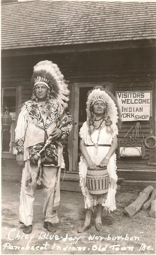 A grayscale image of two people in Native American regalia, include Plains-style war bonnets, stand in front of a shop. A sign reads "Visitors Welcome, Indian Work Shop". The man carries a carved rootclub, the woman a barrel basket.