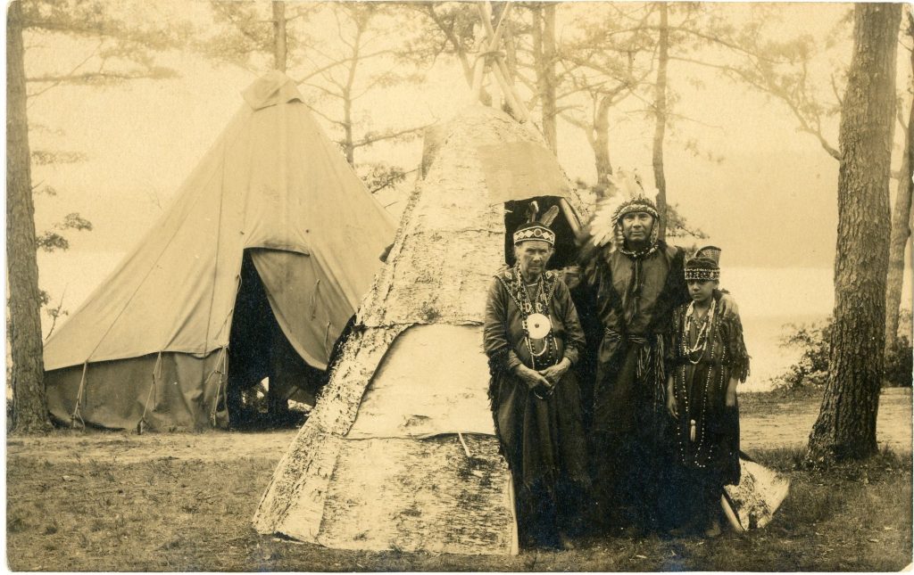 A sepia-tone image of a man, boy, and female elder standing by the entrance of a birchbark tepee. A larger fabric tepee is in the background. The people wear Native dress, including a beaded collar and headband with two feathers and a large silver brooch on the elder, a beaded headband on the boy, and a Plains-style feathered headdress on the man.