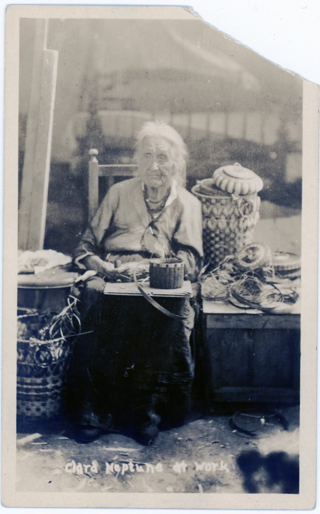 Black and white image of an elderly woman sitting in a museum with an in-progress basket on her lap. She is surrounded by finished baskets.