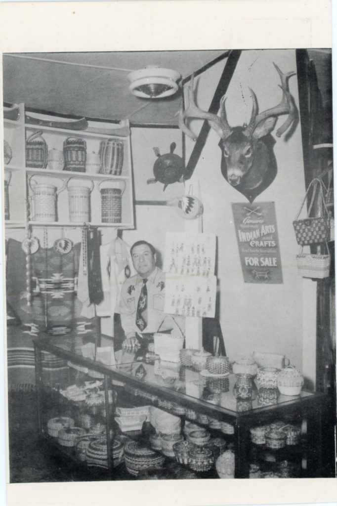 A grayscale image of a man standing behind a glass case. The case and a shelf behind him are crowded with baskets. A poster on the wall behind him reads "Indian Arts and Crafts For Sale".