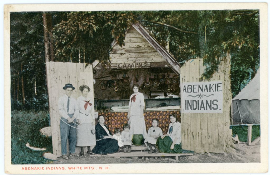 A colorized image of a group of people by a small wooden building, open to display baskets for sale. A sign reads "Abenakie Indians"