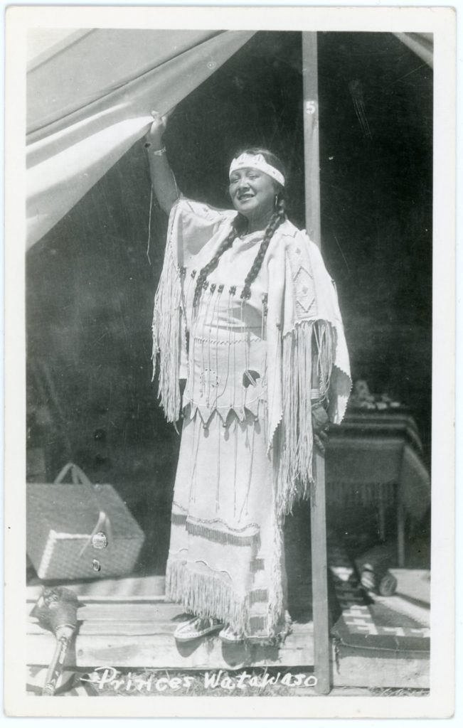 A grayscale image of a smiling woman in Plains-style Native dress standing at the opening of a large tent. Baskets and rootclubs can be seen behind her.