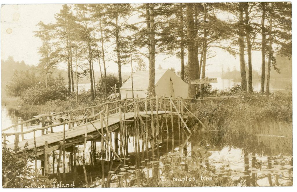 A sepia-tone image of a tent among pine trees on an island reached by a raised wooden boardwalk. A sign by the tent reads "Penobscot Indians Basket Makers"