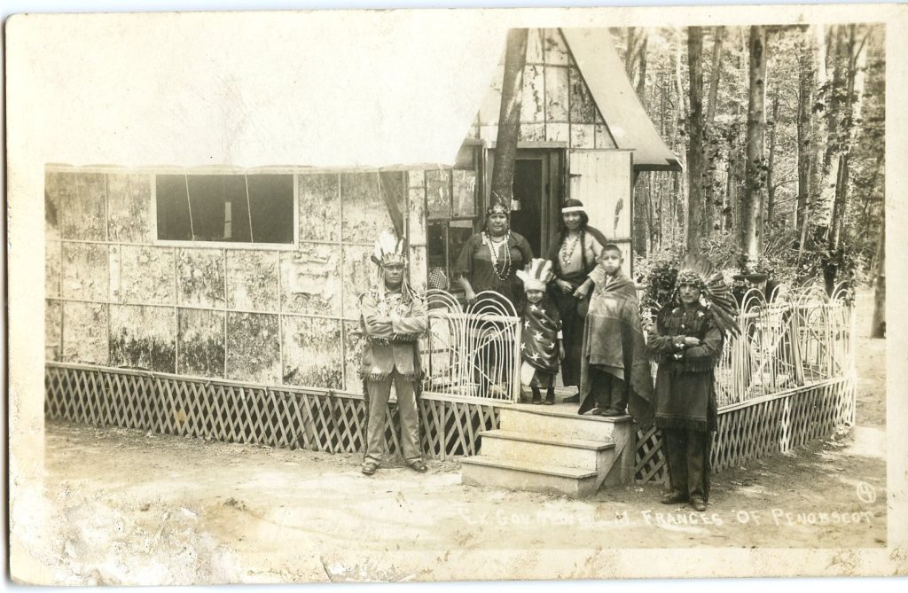 A faded sepia-tone image of a group of people standing on the porch of a house that is sided with birchbark panels.