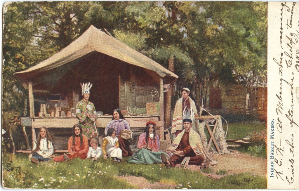 Colorized image of a group of people sitting before a wood and canvas structure. Two wear blankets Plains-style. Two have Wabanaki-style feathered headdresses. One woman has a large bundle of brown ash weavers around her arm. Baskets can be seen on the porch behind the group.