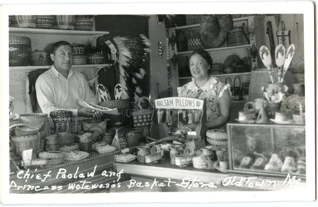 Grayscale image of two people standing behind a store counter. The counter and the shelves behind the people are crowded with baskets, birchbark objects, and other items. A Plains-style war bonnet hangs on the wall between them.