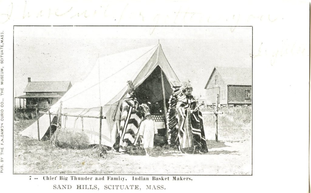 Faded black and white image of three people standing by a canvas tent. Two buildings can be seen in the background. Two men wear blankets and one has a Plains-style war bonnet. The other and a little boy both wear Wabanaki-style feathered headdresses.