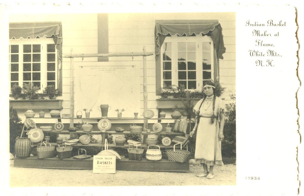 Sepia-tone image of a woman wearing Plains-style dress and braids standing by a display of baskets. A sign reads "Genuine Indian Baskets"