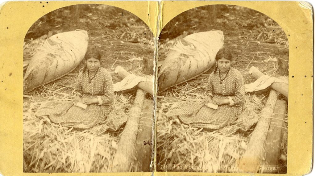 Stereoview image of a young woman with dark hair in Western dress sitting on the ground working on a partially-finished basket. She sits between an upturned birchbark canoe and a small pile of logs. She is surrounded by wood shavings and brown ash splints.