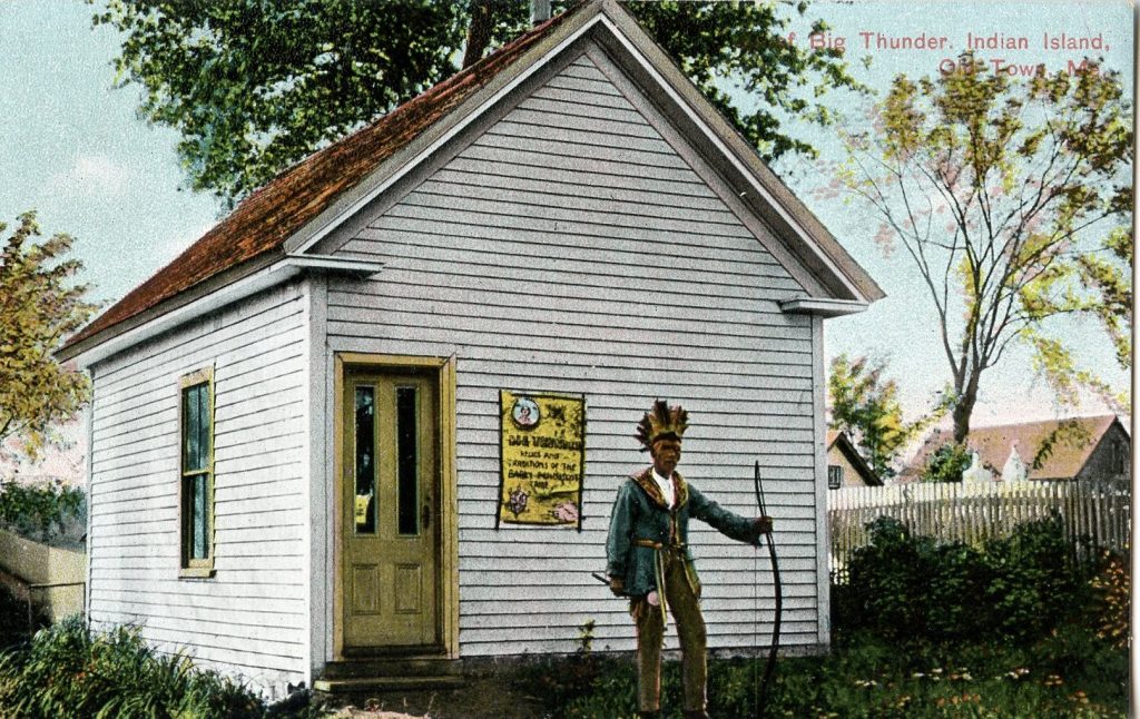 A colorized image of a Penobscot elder standing in front of a small white building. A sign behind him is not legible. He wears traditional dress, including a feathered headdress, and holds a bow in his left hand.