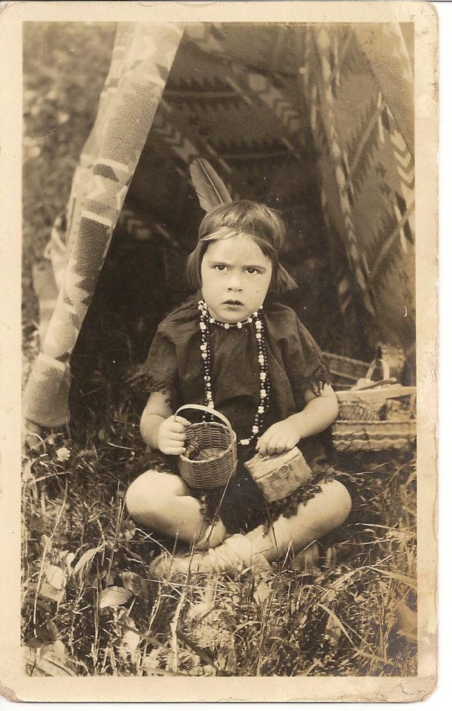 A sepia image of a little girl siting at the opening of a tepee. She is wearing a dark dress, beaded necklace, and single feather behind her head. She is holding two small baskets.