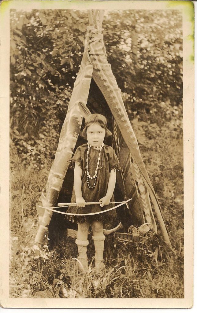 A sepia image of a little girl standing at the opening of a tiny tepee. She is wearing a dark dress, beaded necklace, and single feather behind her head. She is holding a bow.