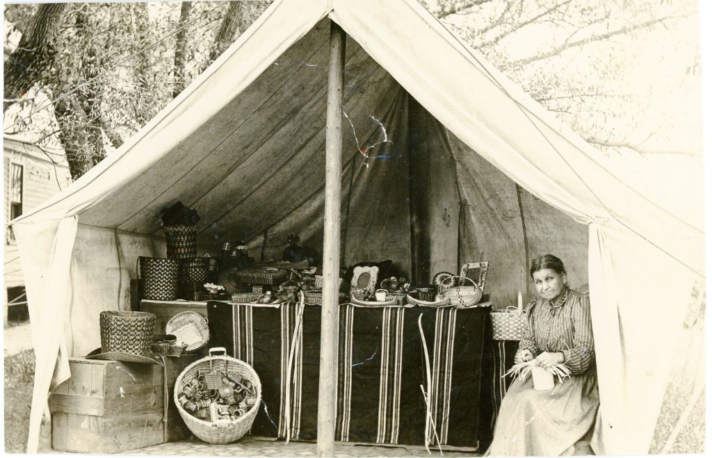 A sepia-tone image of an open canvas tent. Inside is a table covered by a striped blanket on which is piled various baskets and birchbark items. A woman sits to the right, holding a partially-woven basket.