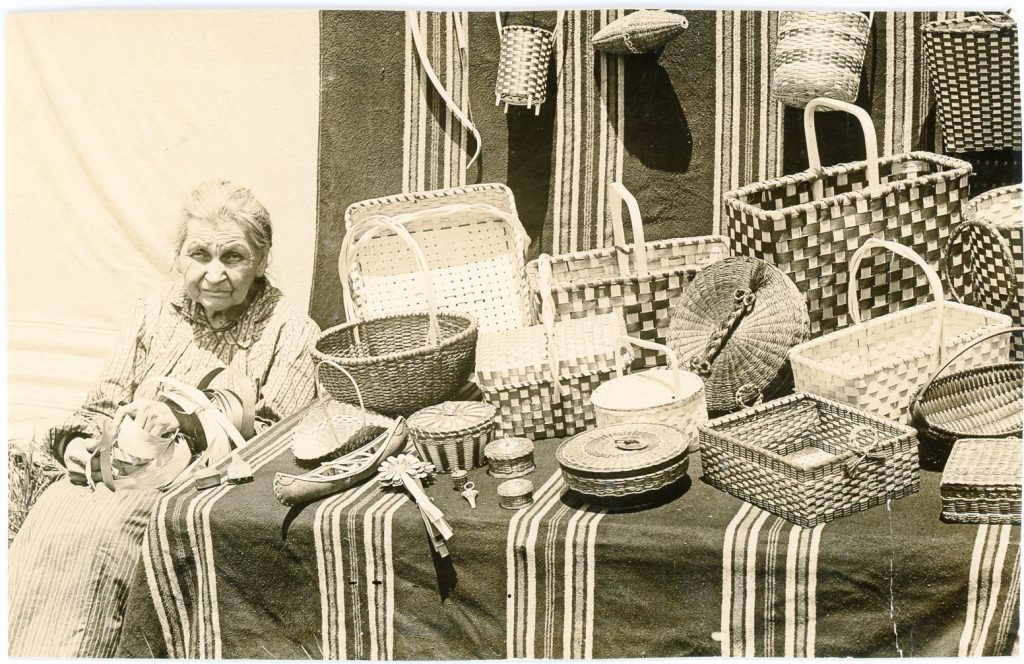 A sepia-tone image of a female elder sitting next to a table covered with a striped cloth and man various baskets. She holds coils of brown ash strips.