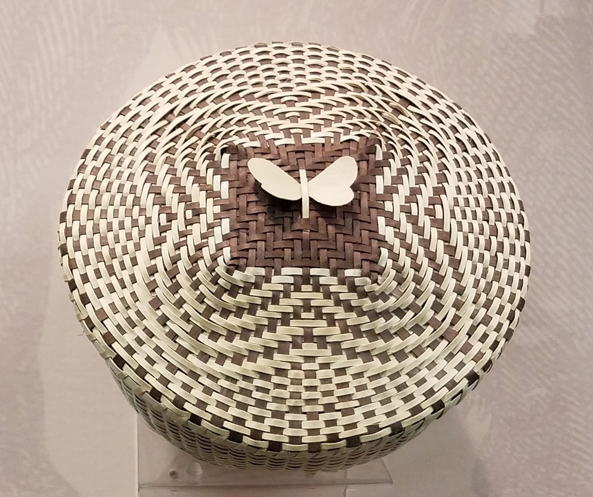 Image of the top of a basket. In the center is a small, simple butterfly made of thin pieces of undyed wood framed against a square of brown at the crown of the basket's cover.