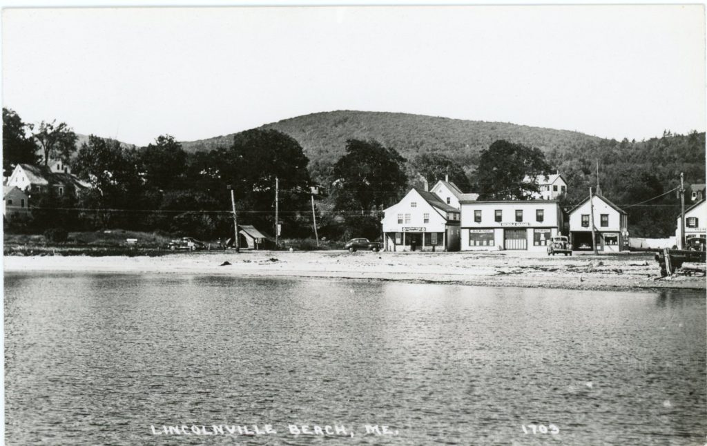 A grayscale image of buildings along a beach taken from the water. In the center can be seen a canvas tent.