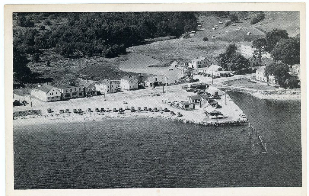 Grayscale, aerial image of a beach with a row of buildings next to a road behind it. A tent can be seen at the far left.