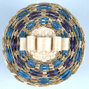 A top-down image of a basket cover with large curls in the middle and sweetgrass braids and brown ash in shades of blue around the edge.