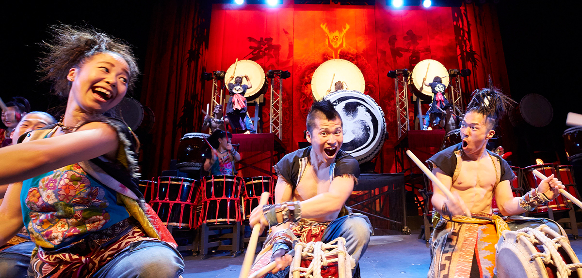 Image of stage performance. Three people are in foreground, singing and moving energetically. In the background multiple levels of drums are played by other people.