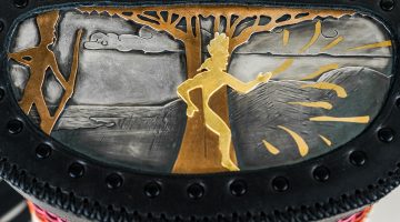 Detail of the top of a basket featuring an image made in metals. A man with a bow on the left, a man coming from a tree center.