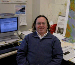 Image of Caucasian man in collared shirt and fleece with shoulder-length hair sitting with desk in background.