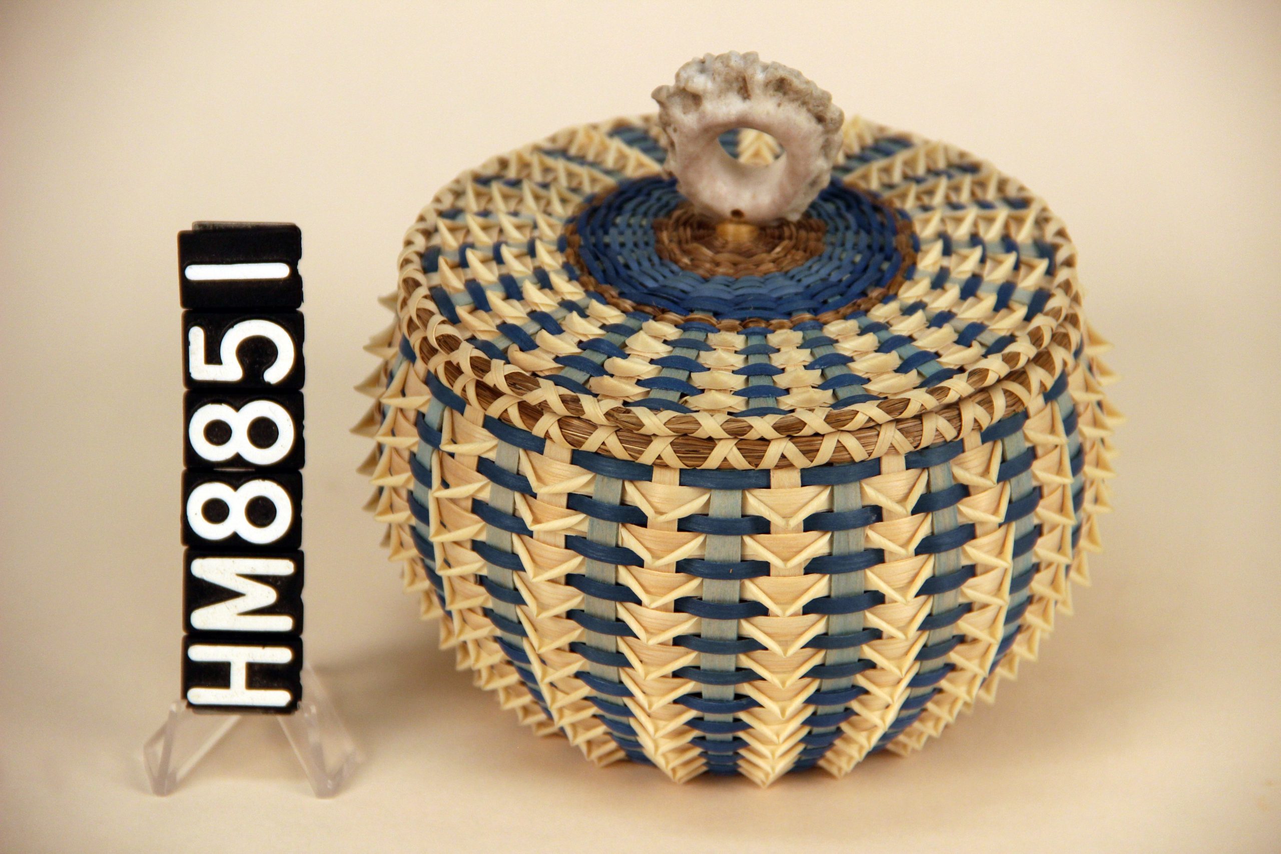 Image of a basket with cover. Basket is intricate with blue weavers and pointed curls.