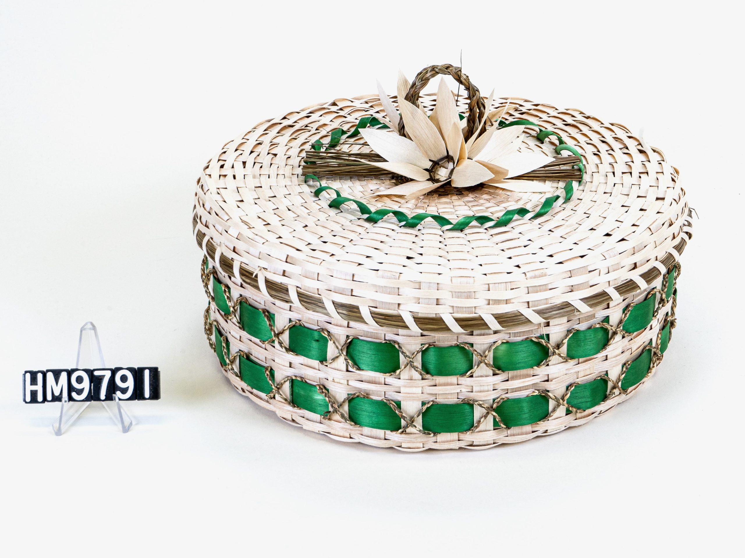 Image of basket, circular, with pale weavers, bright green elements, and details of braided grass. Flowers clustered on cover.