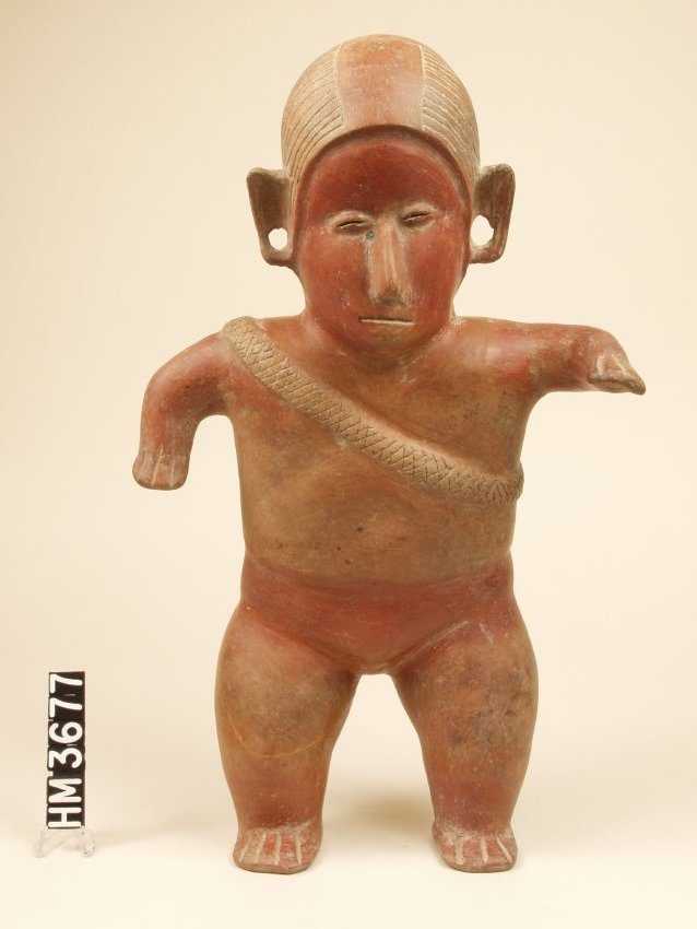Standing ceramic male figure colored red. Large head with incised lines indicating hair. Rectangular ears with holes in them. Coffee-bean eyes, long nose, carved mouth. Arms short and stocky with carved fingers, held out to sides with the right bent at 90 degrees down, and the left bent at 90 degrees and held out front. Conch shell trumpet with lines and circles carved onto ridge strapped on to back by a strap that goes over right shoulder and under left arm. Strap has checkerboard pattern carved into it. Slight indent in abdomen. Thick legs bent slightly with wide feet and long, carved toes. 