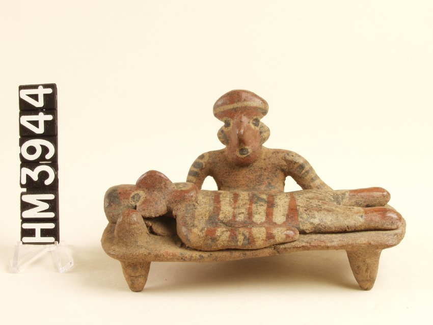 Tiny ceramic object depicting a body laying on a bed supported by four feet. Individual's eyes and mouthy are painted and appear open. A second figure sits to the side. Seated individual has similar facial expression, also painted. 